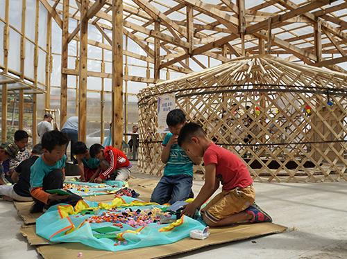 一群孩子坐在木框架和玻璃覆盖结构的地板上. 孩子们正在与五颜六色的材料互动.