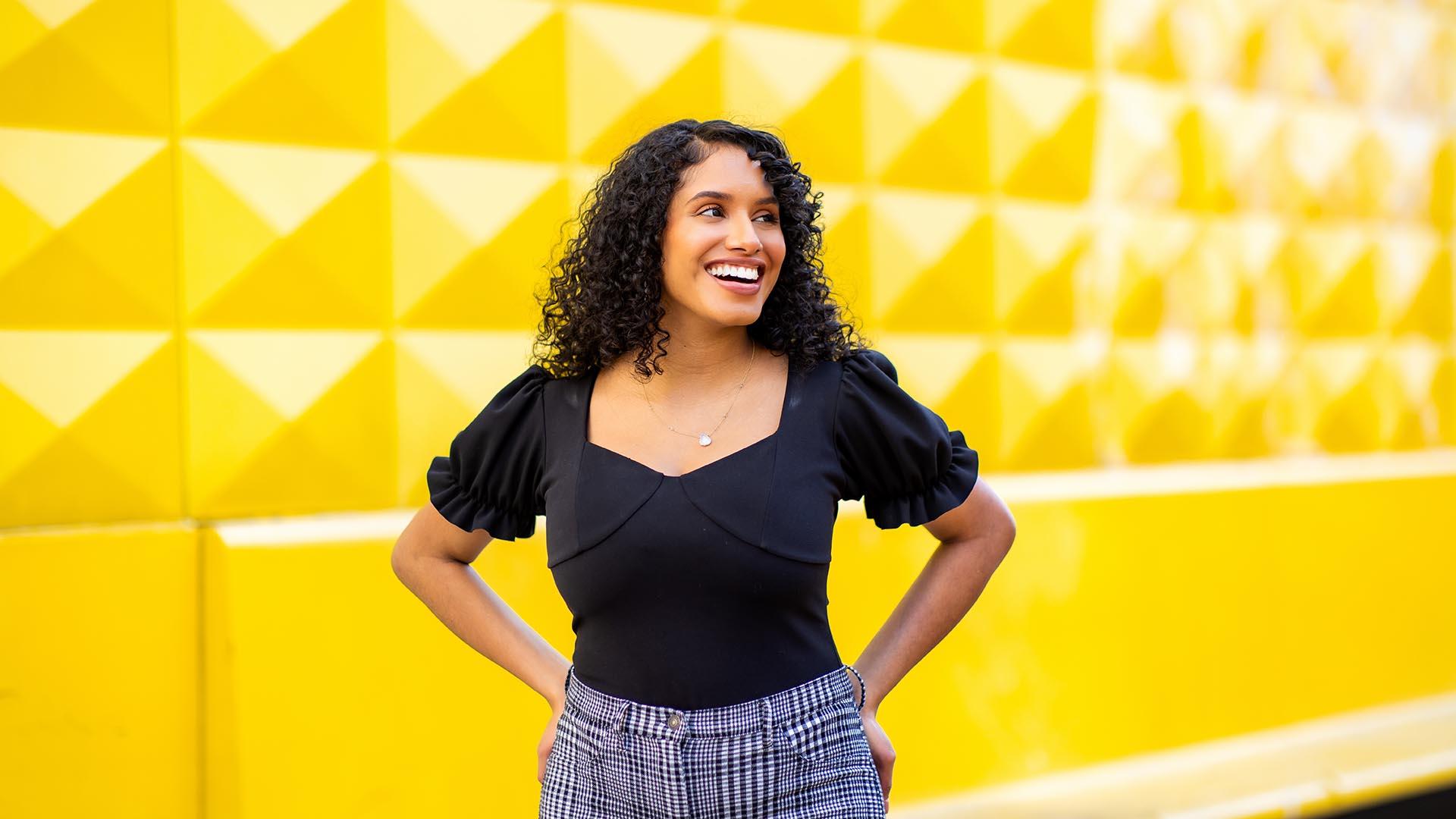 Ines Calvete Barrio in front of a bright yellow wall, her hands on her hips and something happening off to the left that must be making her very happy, evidenced by a glowing smile.